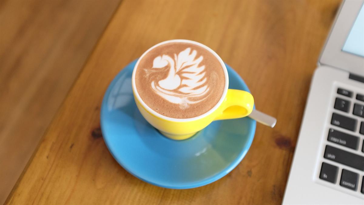 Cangkir berisi kopi dan laptop terbuka di atas meja kayu cafe